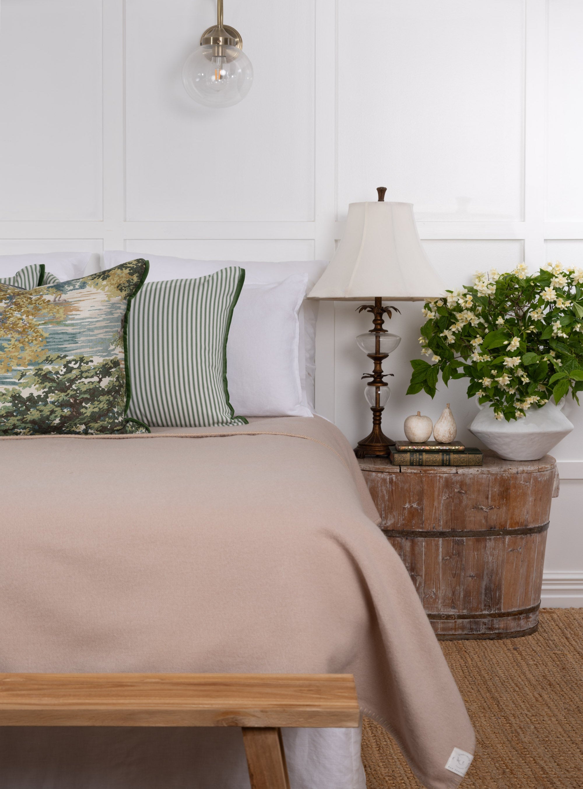 A champagne-colored NZ wool blanket draped over the edge of a bed, with a lamp and decorative pillows in the background.