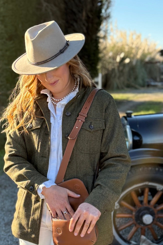 Woman in a green jacket and white pants standing outdoors with a hat and brown bag.