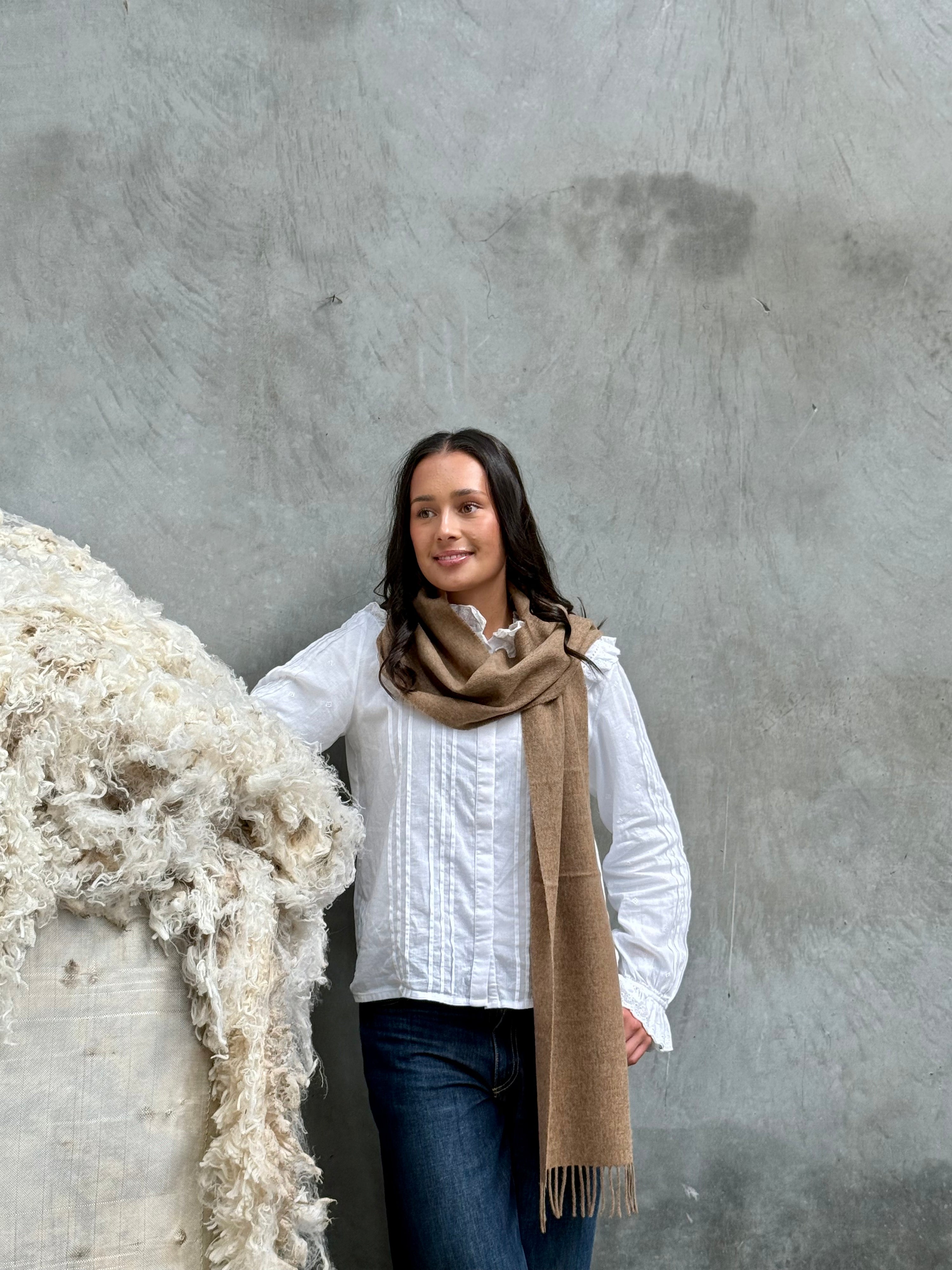 A person standing next to a large pile of wool, wearing a light brown merino scarf with a classic twill weave.